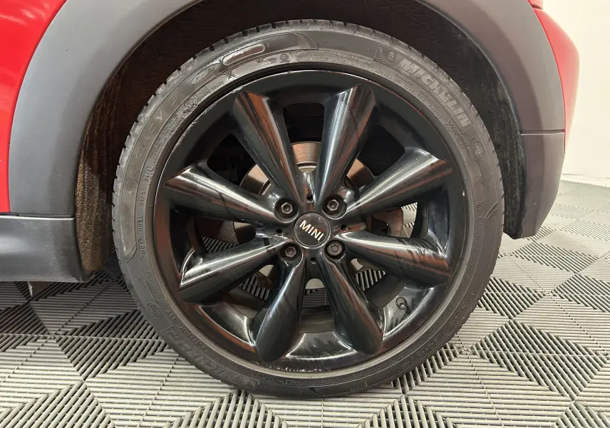 Gros plan sur la roue avant gauche noire brillante d'une MINI Cabriolet rouge, soulignant la jante au design distinctif.