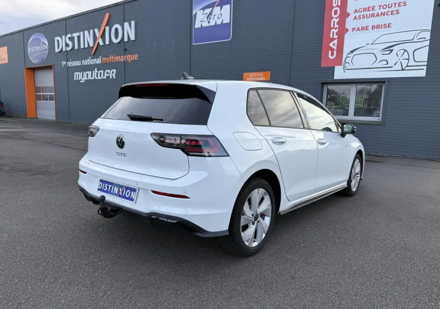 Vue 3/4 arrière droite d'une Volkswagen Golf 1.5 eHybrid blanche nacrée avec attelage et vitres arrière surteintées.