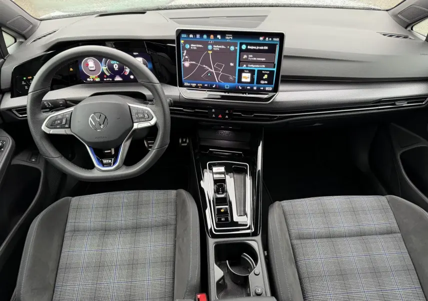 Vue intérieure avant de la Volkswagen Golf 1.5 eHybrid 2025, montrant le volant multifonctions, l’écran tactile central et les sièges sport à motif écossais.