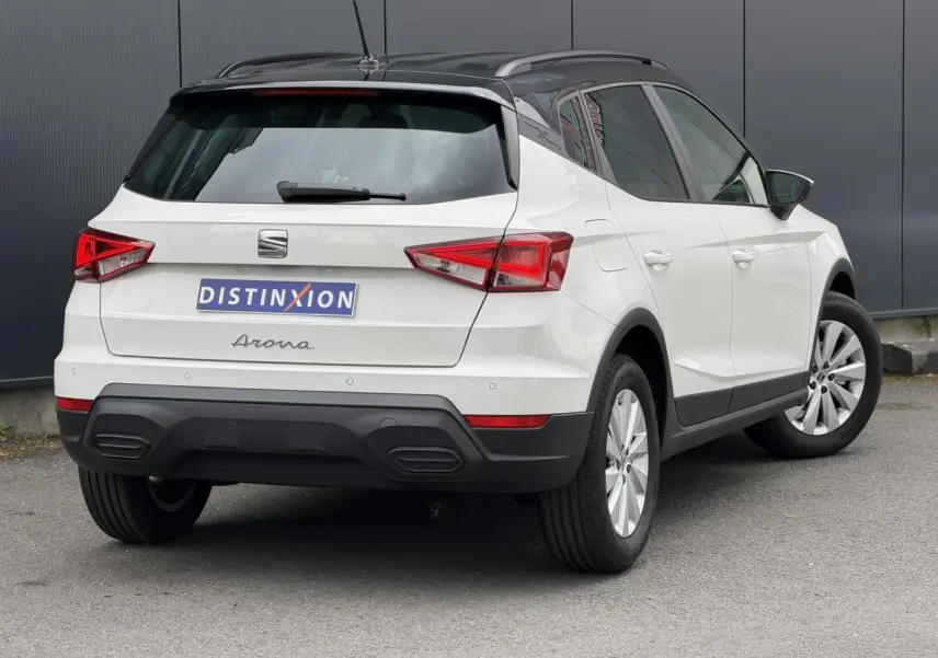 Vue 3/4 arrière droite d'un SEAT Arona blanc avec toit noir et jantes alliage 16 pouces, garé sur sol gris.