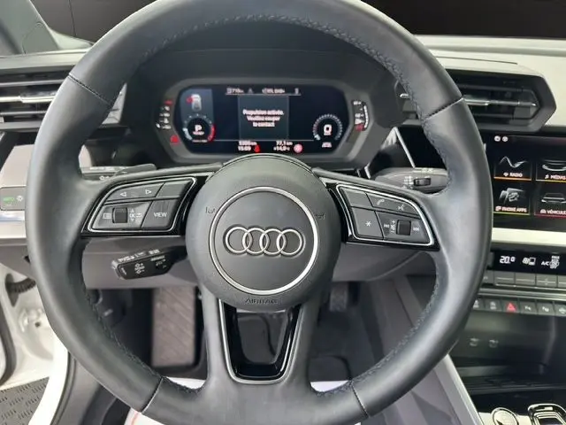 Vue centrée sur le volant cuir noir d'une Audi A3 Sportback blanc Arkona, avec combiné d'instruments digital visible.
