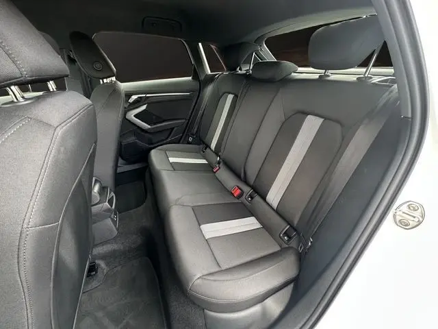 Vue intérieure arrière droite de l'Audi A3 Sportback 2024 blanc Arkona, sièges tissu noir avec bandes grises.