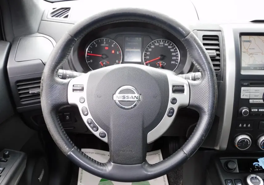 Vue rapprochée du volant Nissan X-TRAIL 2011 avec commandes intégrées et tableau de bord visible en arrière-plan.