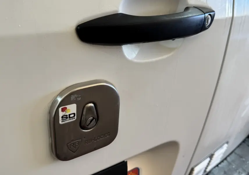 Gros plan sur la serrure antivol argentée et la poignée noire de la porte côté droit du Citroën Berlingo blanc.