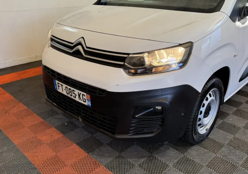 Vue 3/4 avant droit d'un Citroën Berlingo blanc avec pare-chocs noir et phares allumés en intérieur.