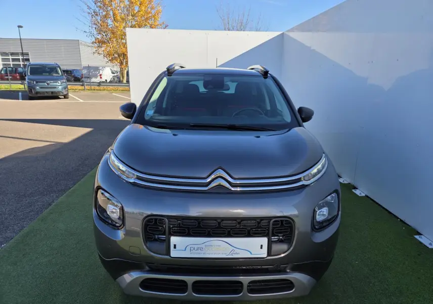 Vue de face d'un Citroën C3 Aircross gris platinium avec toit noir perlé et barres de toit argentées sur fond urbain.
