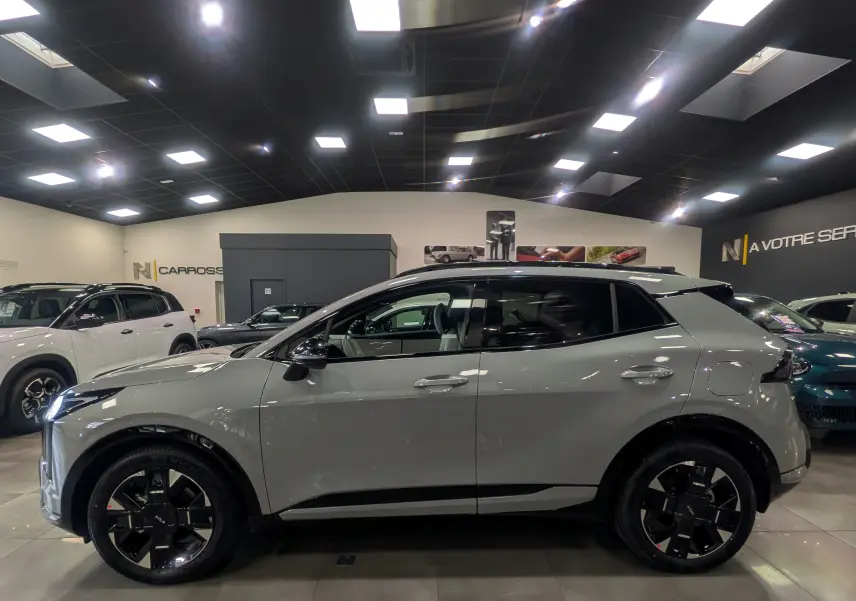 Vue de profil côté gauche d'un KIA SPORTAGE gris céramique avec toit noir, roues noires et intérieur visible en showroom.