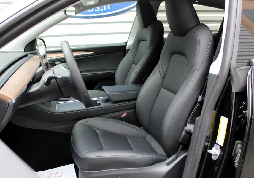 Intérieur de la Tesla Model Y 2024 en cuir noir, vue côté conducteur avec tableau de bord bois et écran central.