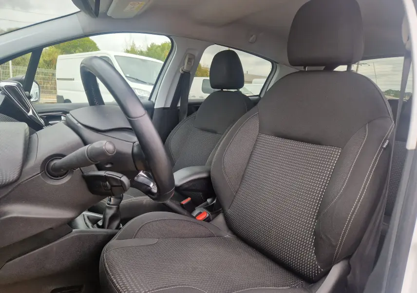 Vue intérieure côté conducteur de la Peugeot 208 blanc nacré, montrant les sièges noirs et le volant avec boîte manuelle.