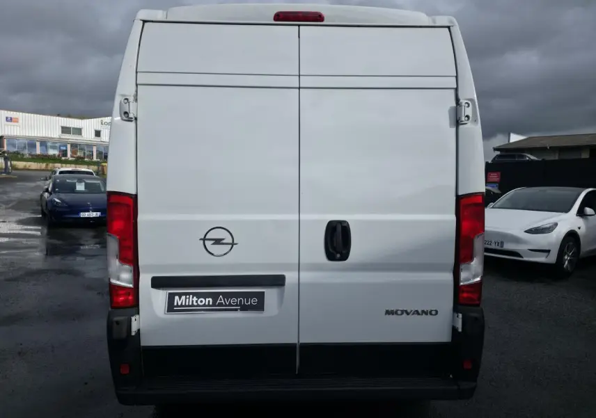 Vue arrière d'un fourgon blanc Opel Movano avec portes battantes et logo visible sous un ciel nuageux.