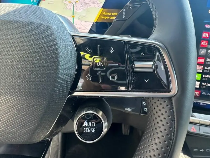 Gros plan sur les commandes gauche du volant noir perforé du Renault Austral 2025 avec bouton Multi-Sense et écran tactile en arrière-plan.