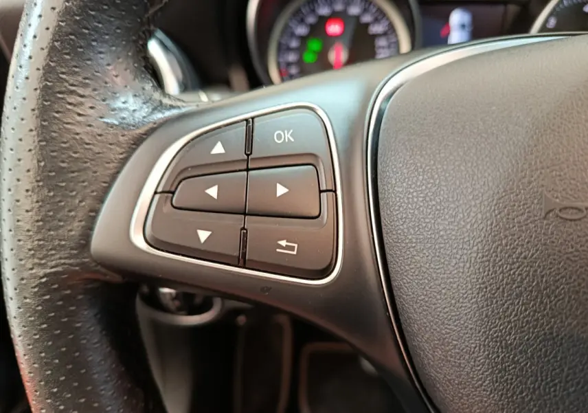 Gros plan sur les commandes gauche du volant en cuir noir du Mercedes GLA 220 d Business Exec 2018, avec tableau de bord flou.