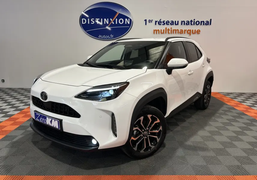 Toyota Yaris Cross blanc en 3/4 avant droit, avec jantes noires et phares LED allumés en showroom.