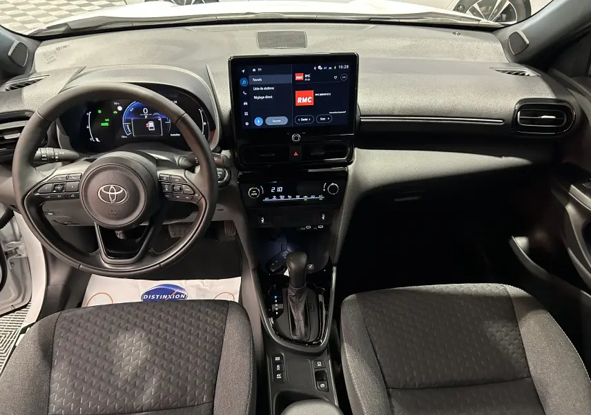 Vue intérieure avant du tableau de bord et des sièges en tissu gris de la Toyota Yaris Cross blanche 2025 avec écran tactile central allumé.