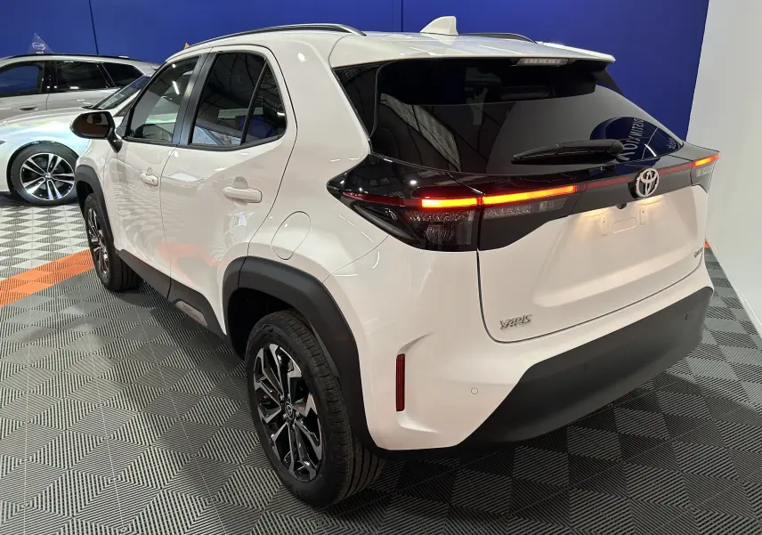 Vue 3/4 arrière droite d'une Toyota Yaris Cross blanche 2025 avec feux arrière LED allumés et jantes noires brillantes.