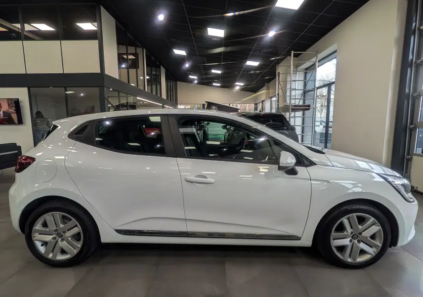 Profil côté gauche d'une Renault Clio V Business blanche Glacier, en intérieur de showroom avec jantes alliage visibles.