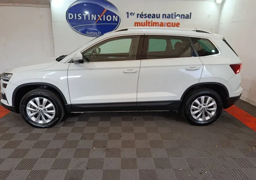 Vue de profil côté gauche d'un SKODA KAROQ blanc 2024, SUV compact avec barres de toit noires et jantes alliage.