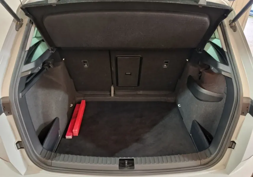 Coffre ouvert vu de l'arrière d'un Skoda Karoq 2024 beige, avec tapis noir et deux objets rouges à gauche.