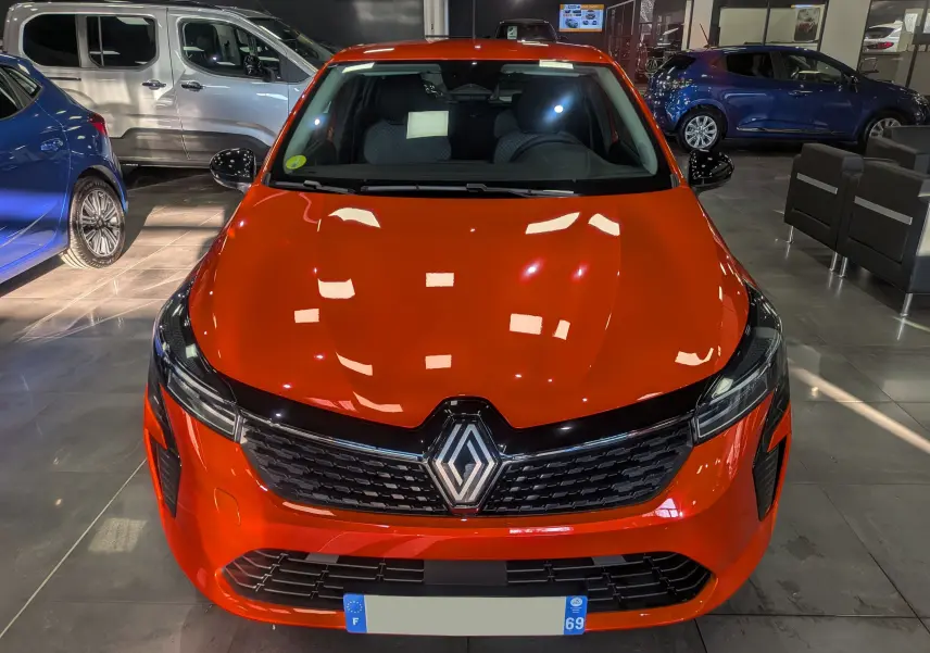 Vue avant d'une Renault Clio V Orange Valencia 2024 en intérieur, avec calandre noire et logo central visible.