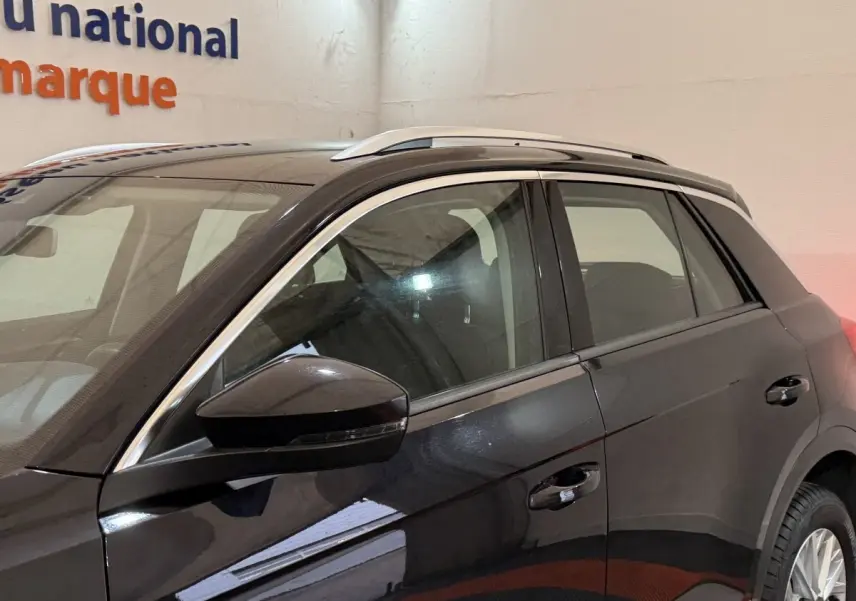 Vue latérale côté gauche d'un Volkswagen T-Roc noir 2020 avec barres de toit argentées en intérieur showroom.
