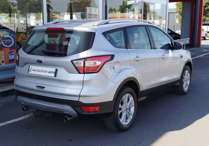 Vue 3/4 arrière droite d'un Ford Kuga gris clair 2019 avec double sortie d'échappement et barres de toit visibles.