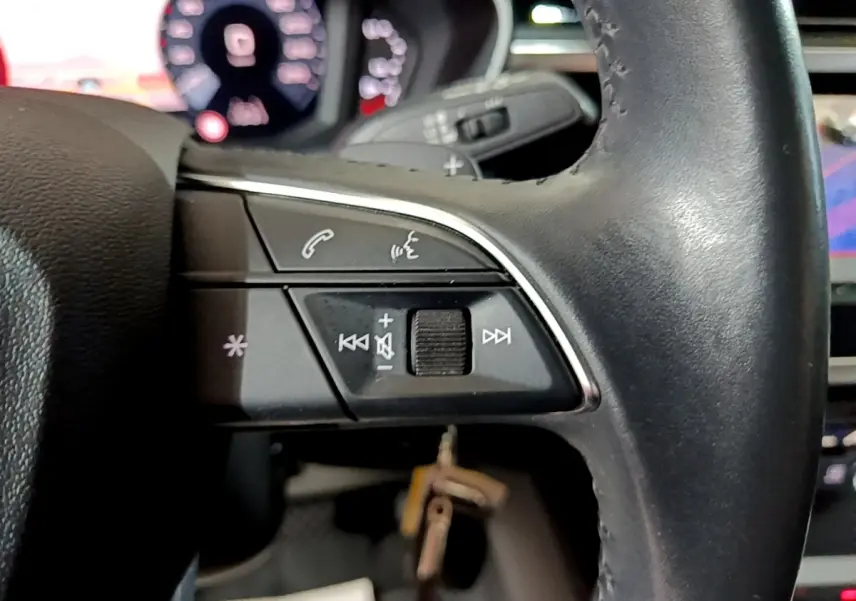 Gros plan sur les commandes du volant cuir noir de l'Audi Q3 35 TDI 150 S tronic Business Line, avec tableau de bord numérique flou en arrière-plan.