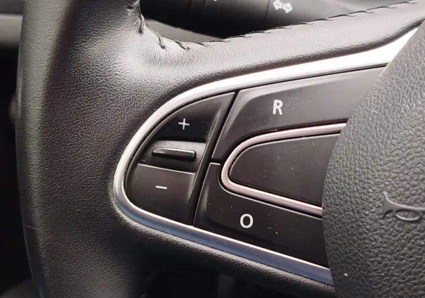 Gros plan sur les commandes noires du volant en cuir de la Renault Mégane gris Titanium, avec boutons +, -, R et O visibles.