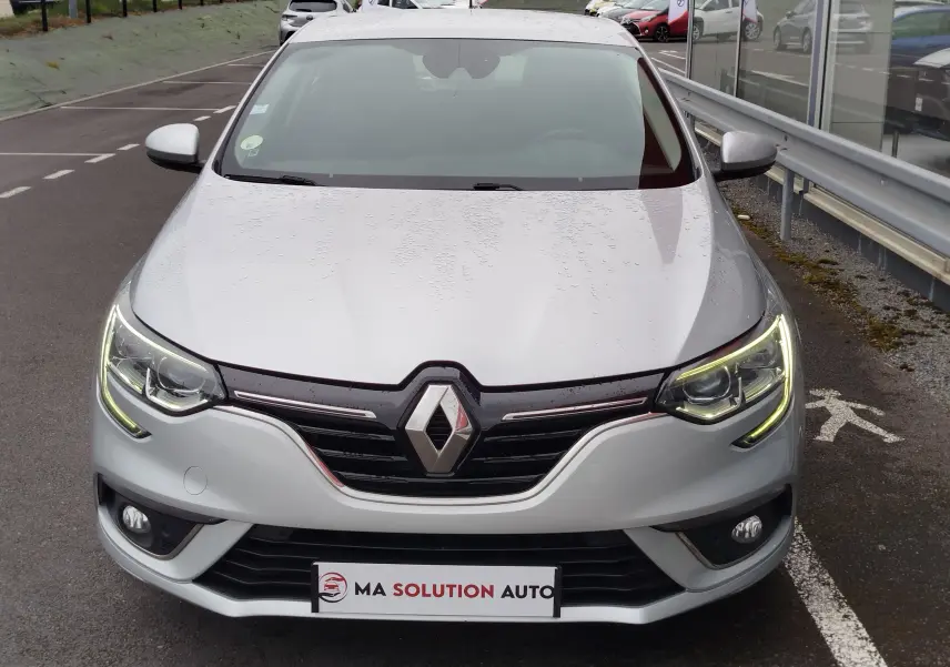 Vue frontale d'une Renault Mégane gris titanium avec phares allumés et logo Renault bien visible sur la calandre.
