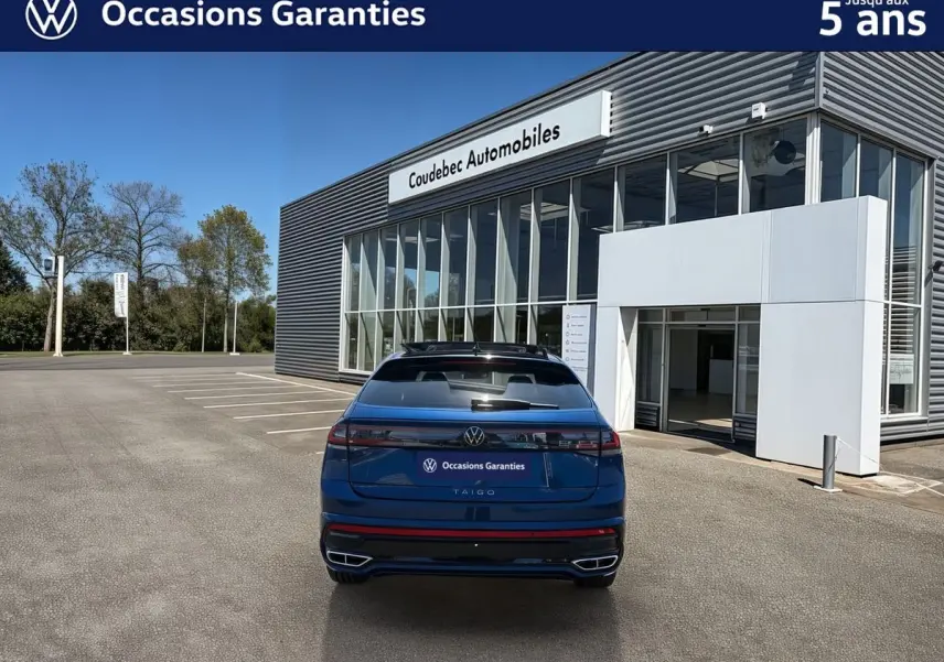 Vue arrière d’un Volkswagen Taigo bleu métallisé 2025 avec toit ouvrant électrique, stationné devant un bâtiment moderne.