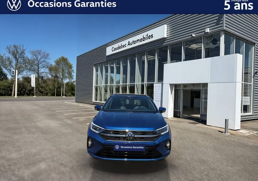 Vue frontale d'un Volkswagen Taigo bleu métallisé garé devant un bâtiment moderne de concession automobile.