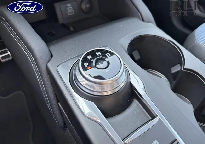 Gros plan sur la molette de boîte automatique centrale du Ford Kuga bleu avec accoudoir noir et porte-gobelets.