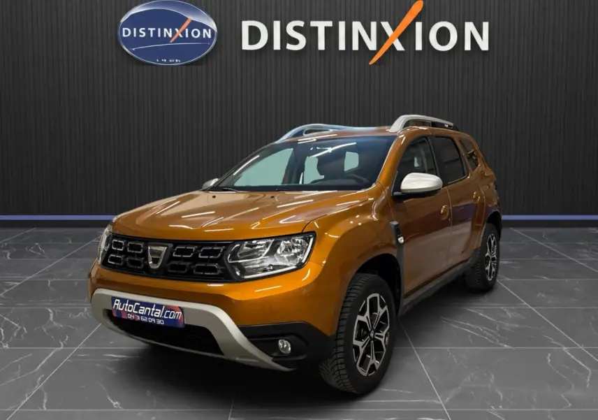Dacia Duster orange en 3/4 avant droit, avec jantes bi-ton et rétroviseurs blancs, sur sol gris intérieur showroom.