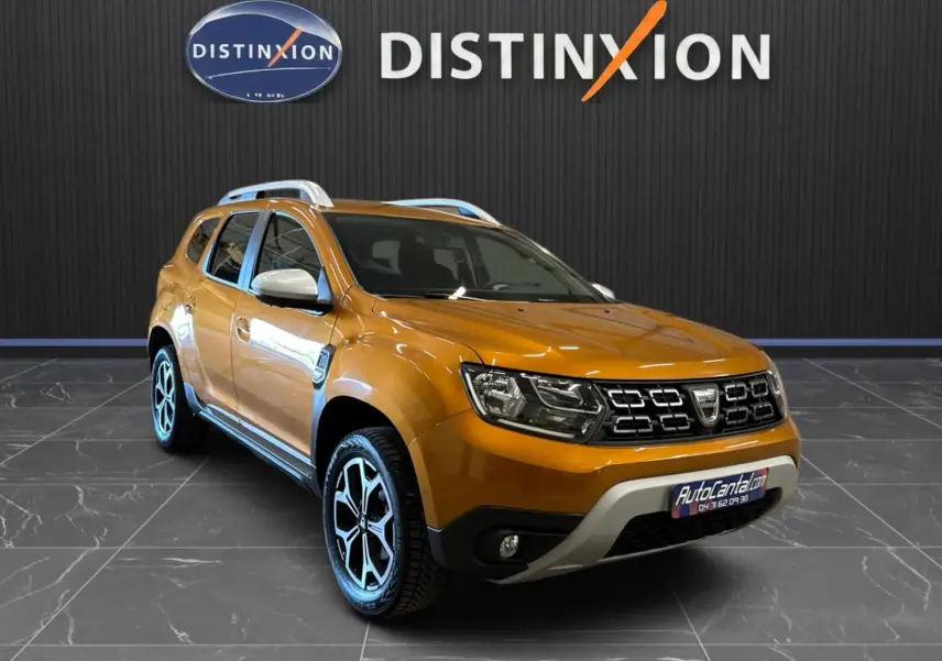 Dacia Duster orange en 3/4 avant droit, avec jantes bi-ton et barres de toit argentées, sur sol carrelé intérieur showroom.