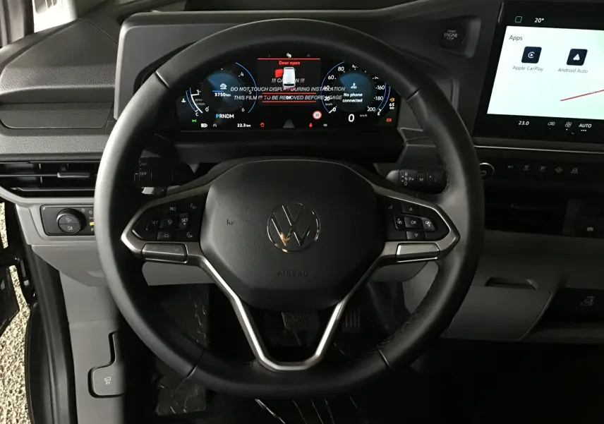 Vue intérieure centrée sur le volant noir du Volkswagen Transporter T7 avec tableau de bord numérique et écran tactile à droite.