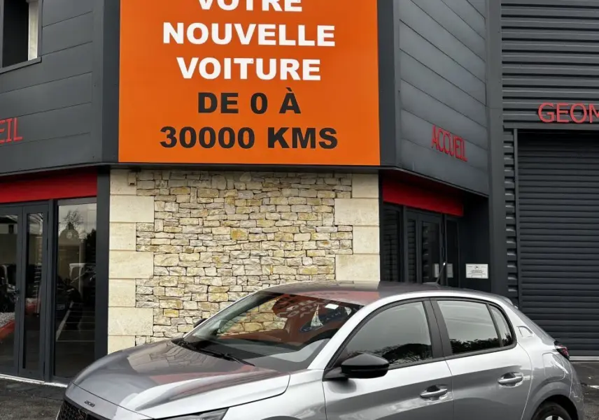Peugeot 208 PureTech 100 S&S Active gris clair vue 3/4 avant droit devant un bâtiment avec panneau orange Distinxion.