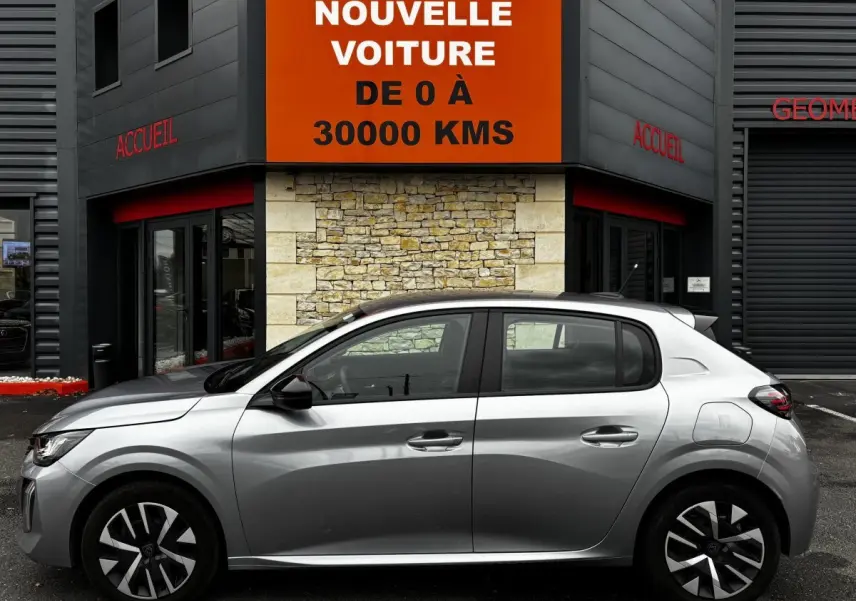 Profil côté gauche d'une Peugeot 208 PureTech 100 S&S Active gris clair, garée devant un bâtiment avec panneau orange.