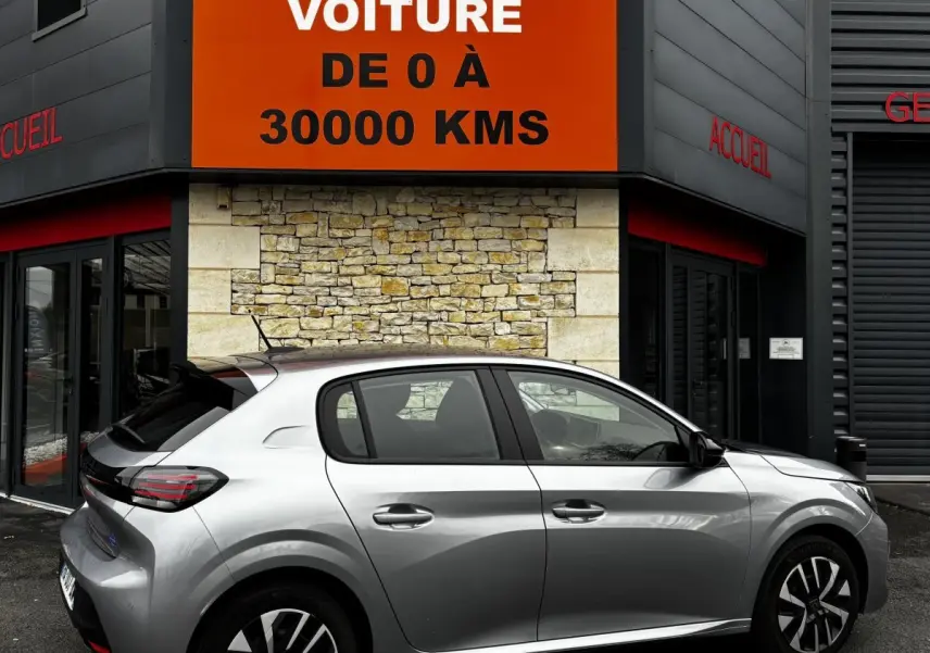 Peugeot 208 gris clair vue de profil côté droit devant un mur en pierre avec panneau orange publicitaire.