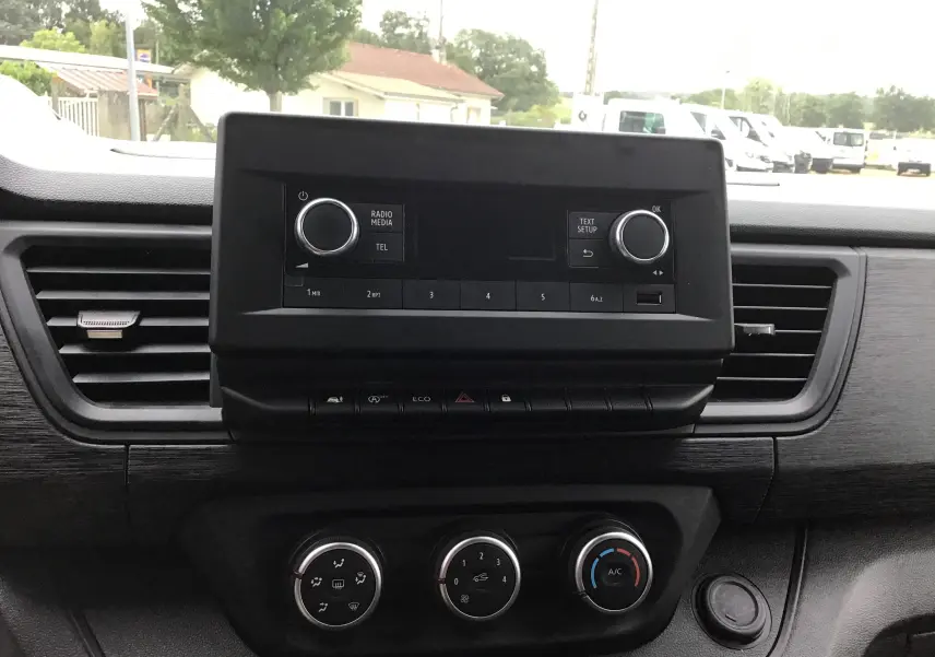 Vue intérieure centrée sur la console centrale noire du Renault Trafic Fourgon blanc, avec commandes radio et climatisation visibles.