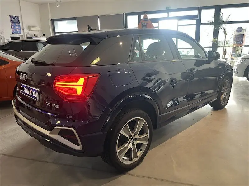 Audi Q2 30 TFSI S-Line bleu Navarre métal vu en 3/4 arrière droit avec feux arrière allumés en intérieur showroom.