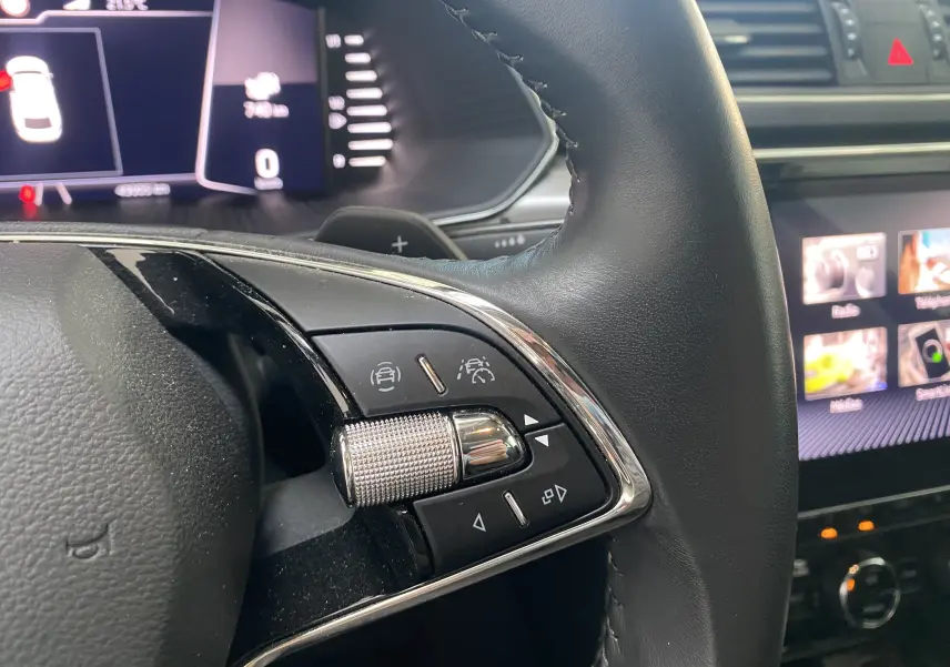 Gros plan sur les commandes du volant en cuir noir d'une Skoda Superb Combi bleu énergie, avec tableau de bord numérique en arrière-plan.