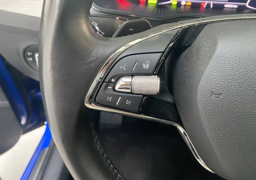Gros plan sur les commandes gauche du volant en cuir noir du Skoda Superb Combi bleu énergie, avec molette chromée et tableau de bord numérique.