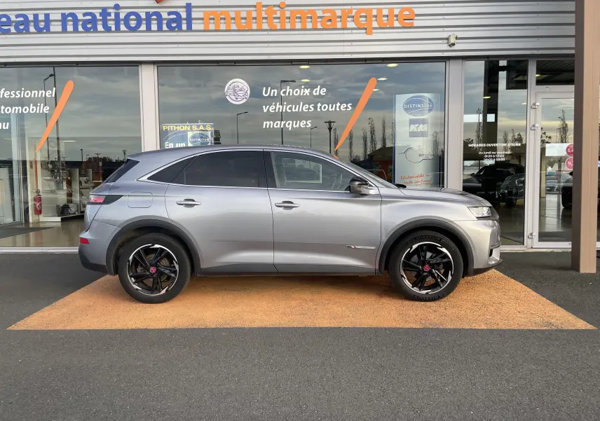 Profil côté gauche d'un DS7 gris 2021 avec jantes noires bicolores, garé devant une vitrine multimarque.
