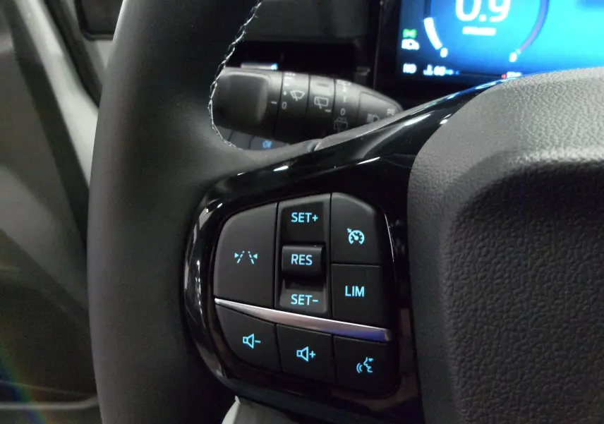 Gros plan sur les commandes au volant gauche du Ford Puma gris, avec régulateur de vitesse et contrôle audio rétroéclairés.