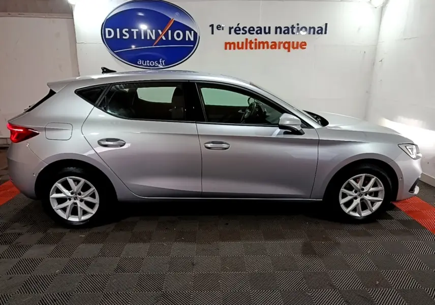 Profil côté droit d'une SEAT LEON gris clair 2023 dans un showroom avec logo Distinxion en arrière-plan.