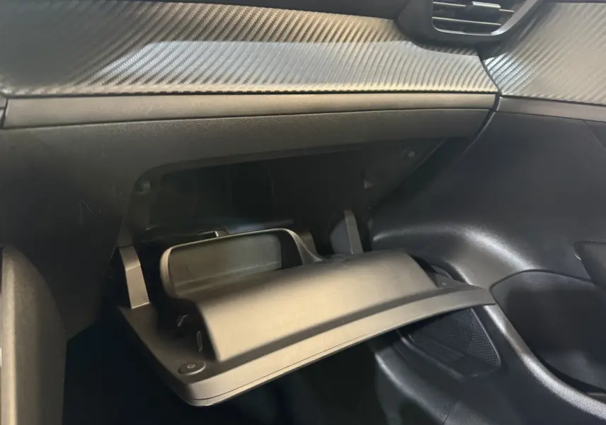 Intérieur de la Peugeot 208 PureTech 75 S&S Like 2020, vue sur la boîte à gants ouverte avec finition noire et effet carbone.