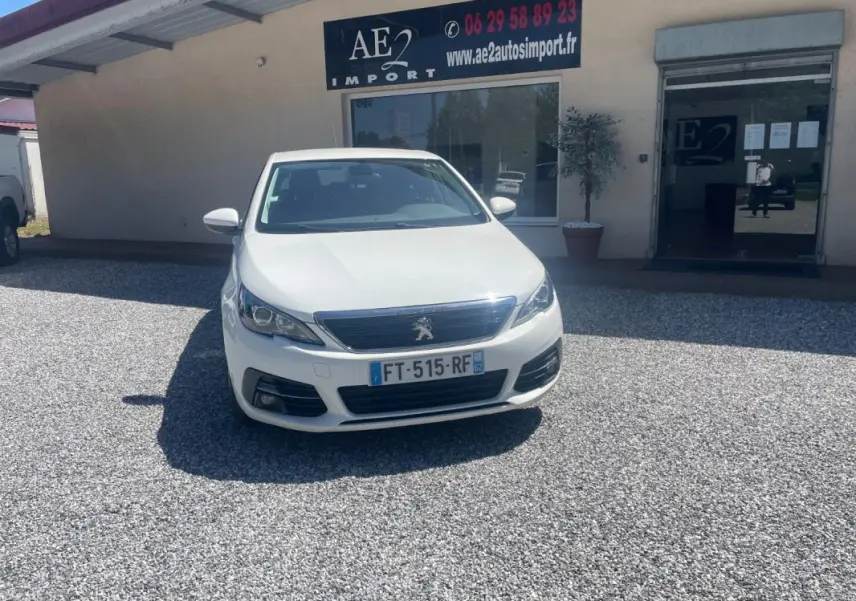 Peugeot 308 blanc vue de face, stationnée devant un bâtiment avec panneau AE2 Import visible en arrière-plan.