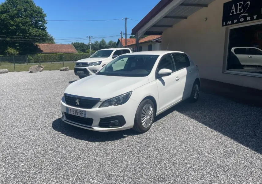 Peugeot 308 blanc vue 3/4 avant droit, garée sur un sol gravillonné devant un bâtiment avec enseigne AE2 Import.