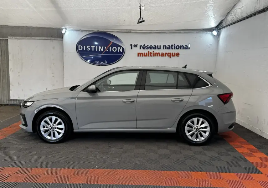 Vue de profil côté gauche d'une Skoda Scala gris clair 2025 dans un showroom avec sol en damier noir et orange.
