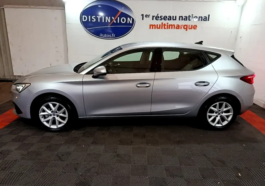 Profil côté gauche d'une SEAT Leon gris clair 2023, en intérieur sur sol damier, avec jantes alliage et feux arrière visibles.