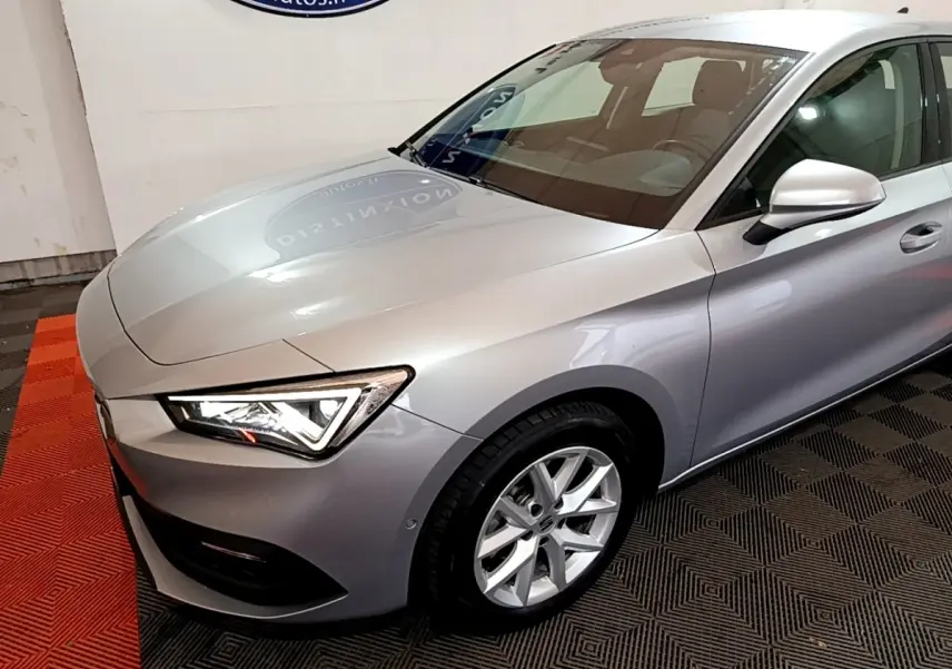 Vue 3/4 avant droit d'une SEAT Leon gris clair avec phares LED et jantes alliage en intérieur showroom.